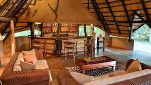 An indoor lounge area with a thatched roof, wooden furniture, and a bar with bottles. - Ivory Lodge (Dete)