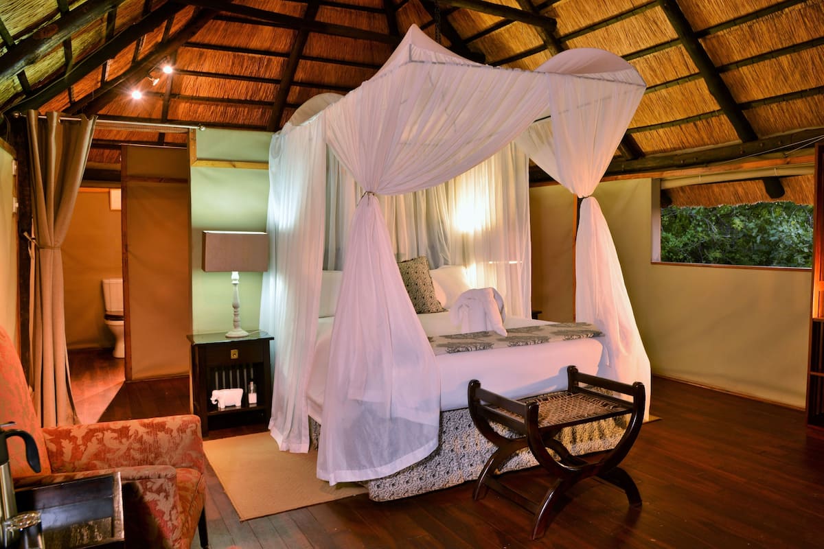 A bedroom with a four-poster bed, a nightstand with a lamp, a chair, and a view of greenery outside.