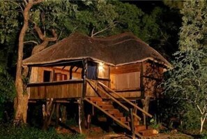 A wooden treehouse with a thatched roof, built on stilts, and accessible by a wooden staircase.