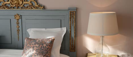 Una cama con sábanas blancas y un cojín floral, un cabecero azul con detalles dorados, y una lámpara de mesa sobre una mesita de noche.