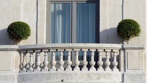 Une façade d’immeuble avec un balcon, une fenêtre ornée de rideaux bleus et deux petits arbustes taillés en forme.