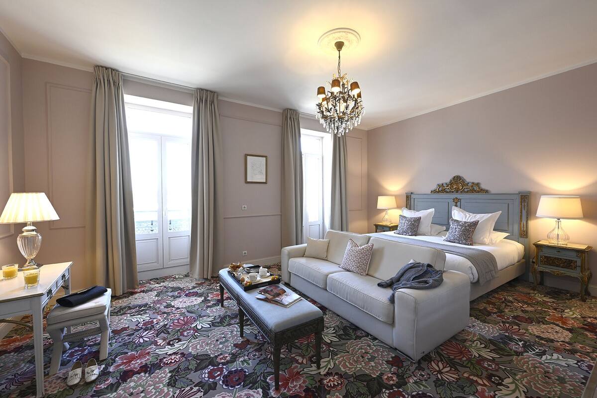 Une chambre d’hôtel avec un canapé blanc, un lit doté d’une tête de lit bleue, un lustre et une moquette à motifs floraux.