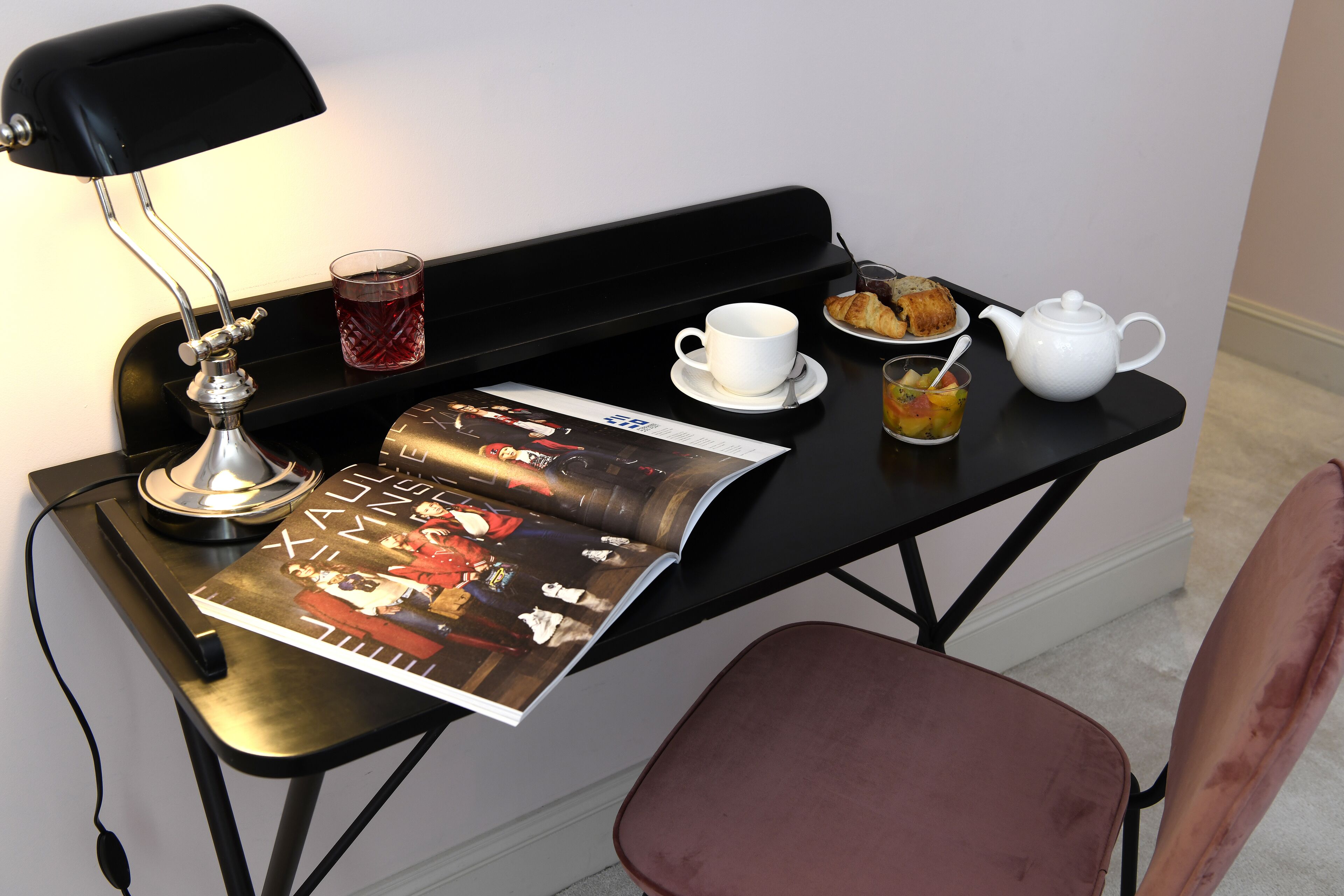 Un pequeño escritorio negro con una revista, una taza, una tetera y un plato de cruasanes.