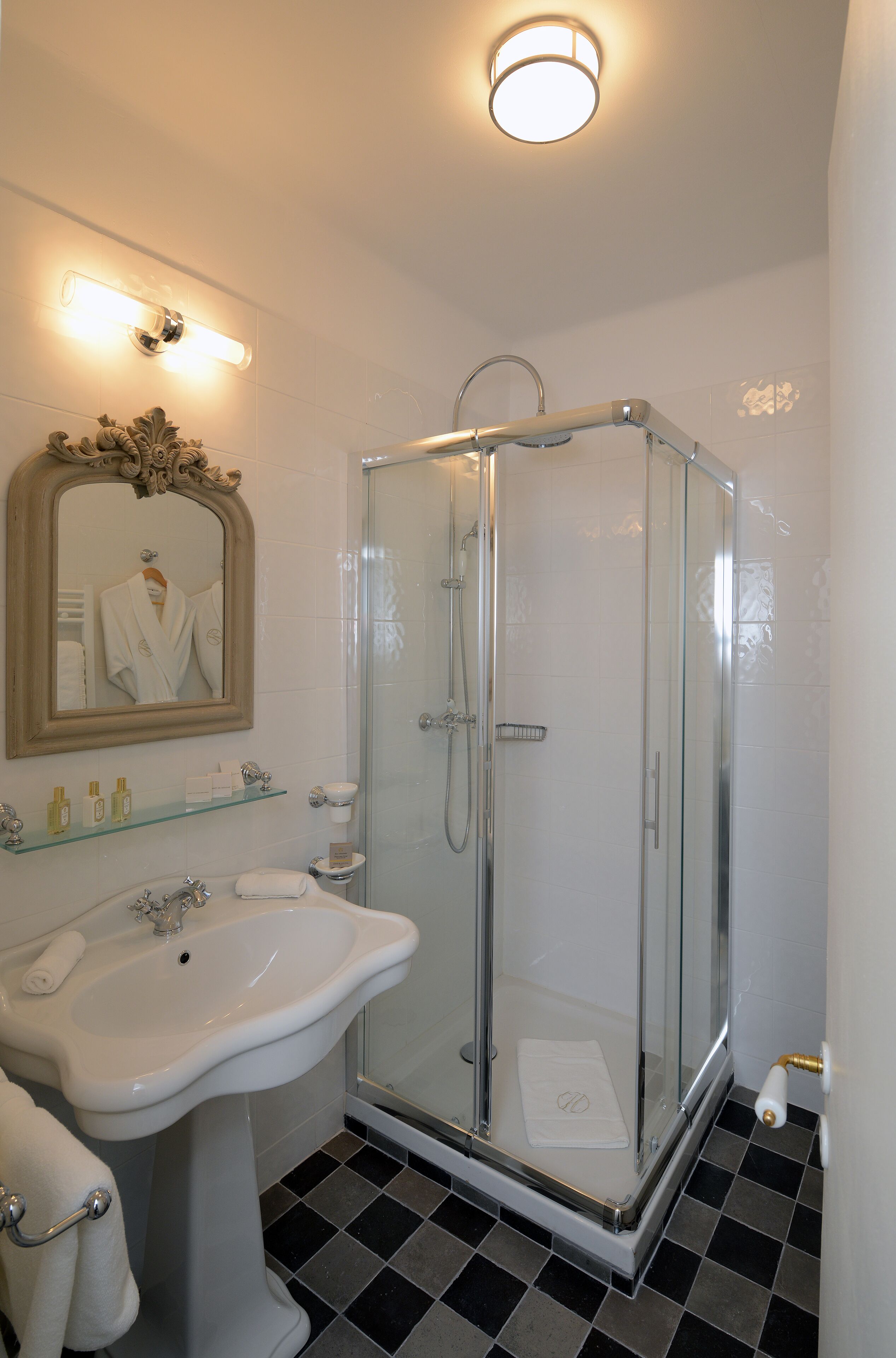 A bathroom with a glass-enclosed shower, a pedestal sink, a mirror with a towel hanging, and a shelf with toiletries.