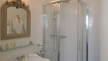 A bathroom with a glass-enclosed shower, a pedestal sink, a mirror with a towel hanging, and a shelf with toiletries.
