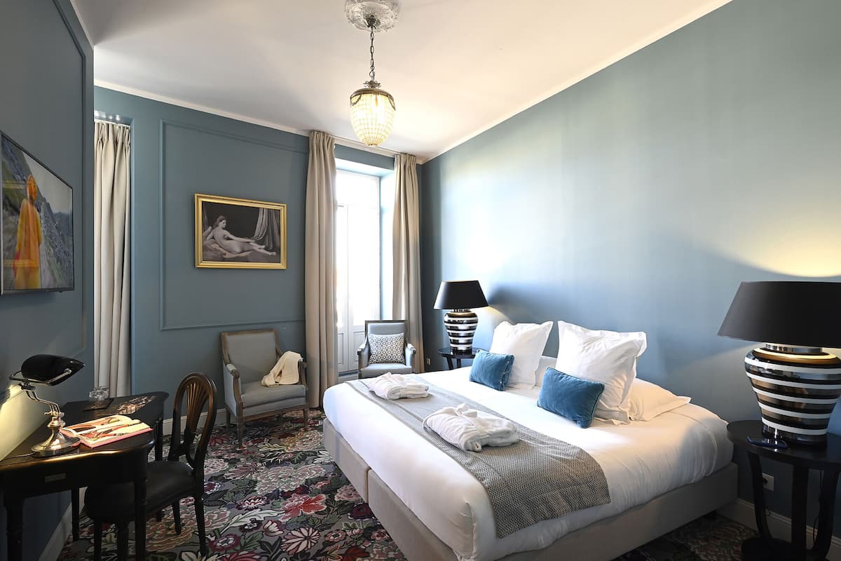 Une chambre d’hôtel avec un grand lit, deux tables de chevet, un bureau avec une chaise, une chaise avec une couverture, un tableau au mur et un lustre.
