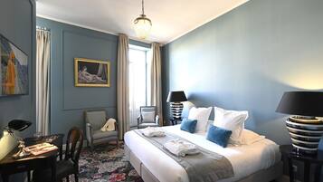 A hotel room with a large bed, two bedside tables, a desk with a chair, a chair with a blanket, a painting on the wall, and a chandelier.
