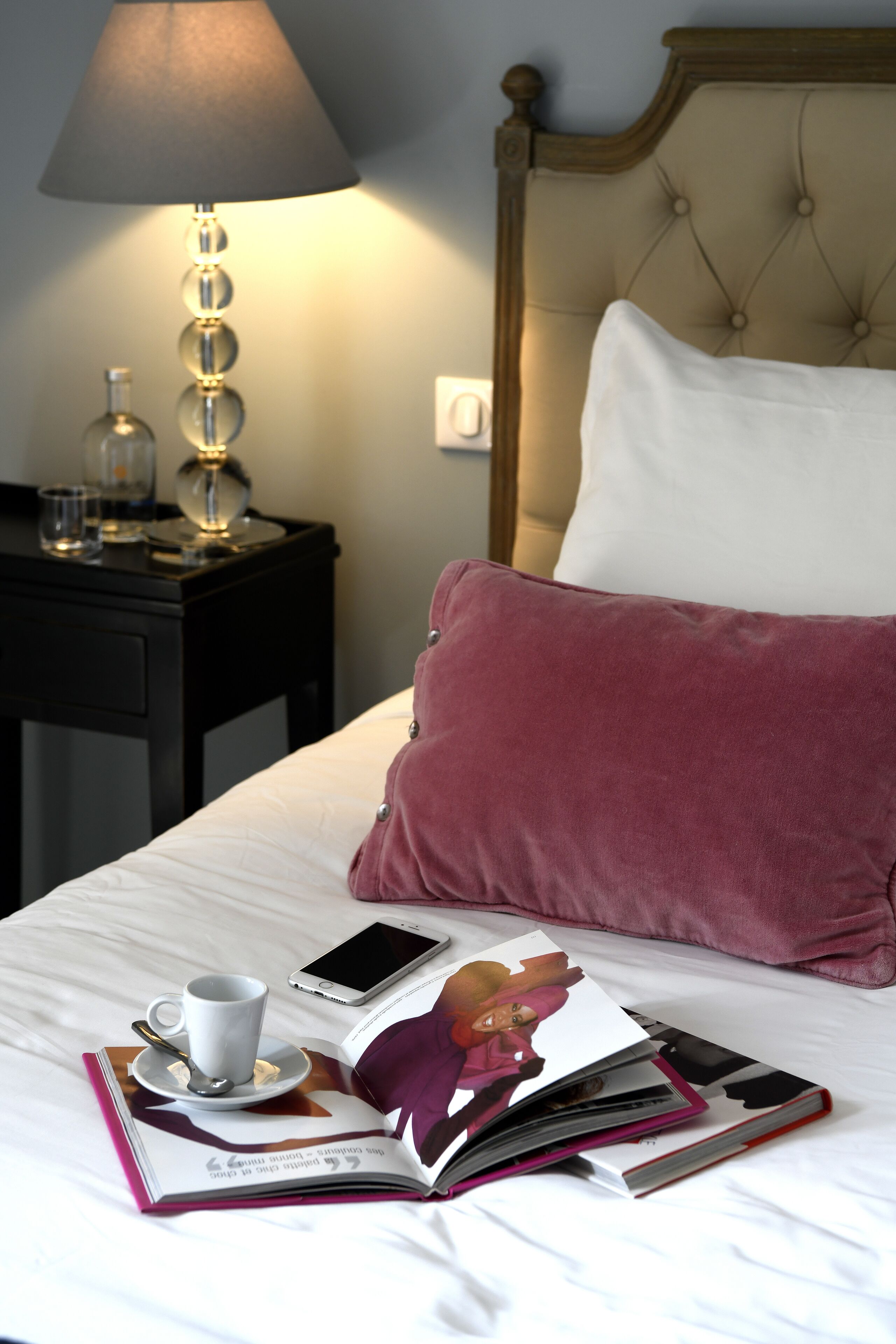 A neatly made bed with a white and pink pillow, a bedside table with a lamp, a book, and a cup with a spoon.