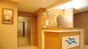 A reception desk with a glass top, wooden paneling, and a logo. There are three clocks above the desk and a framed picture on the wall. - Deniz Palace Hotel (Istanbul)