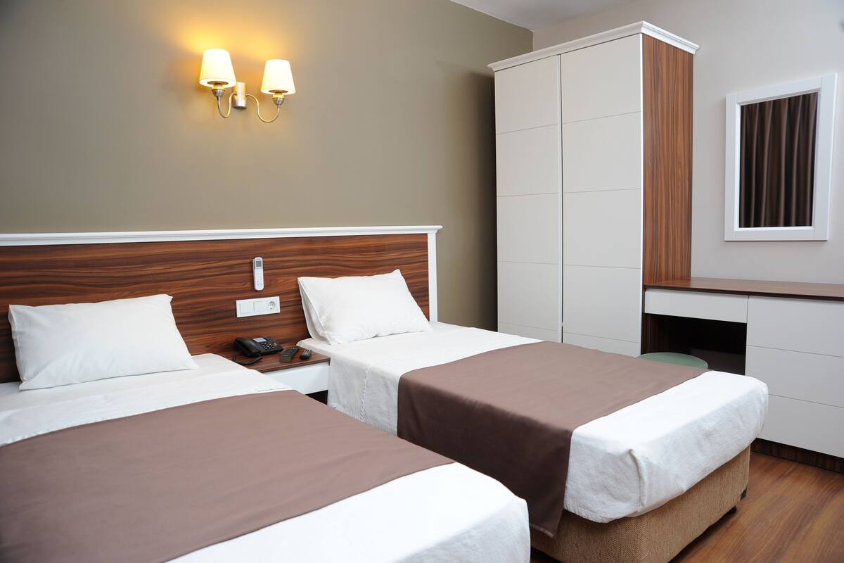 A hotel room with two beds, a wardrobe, a desk, and a wall-mounted lamp.