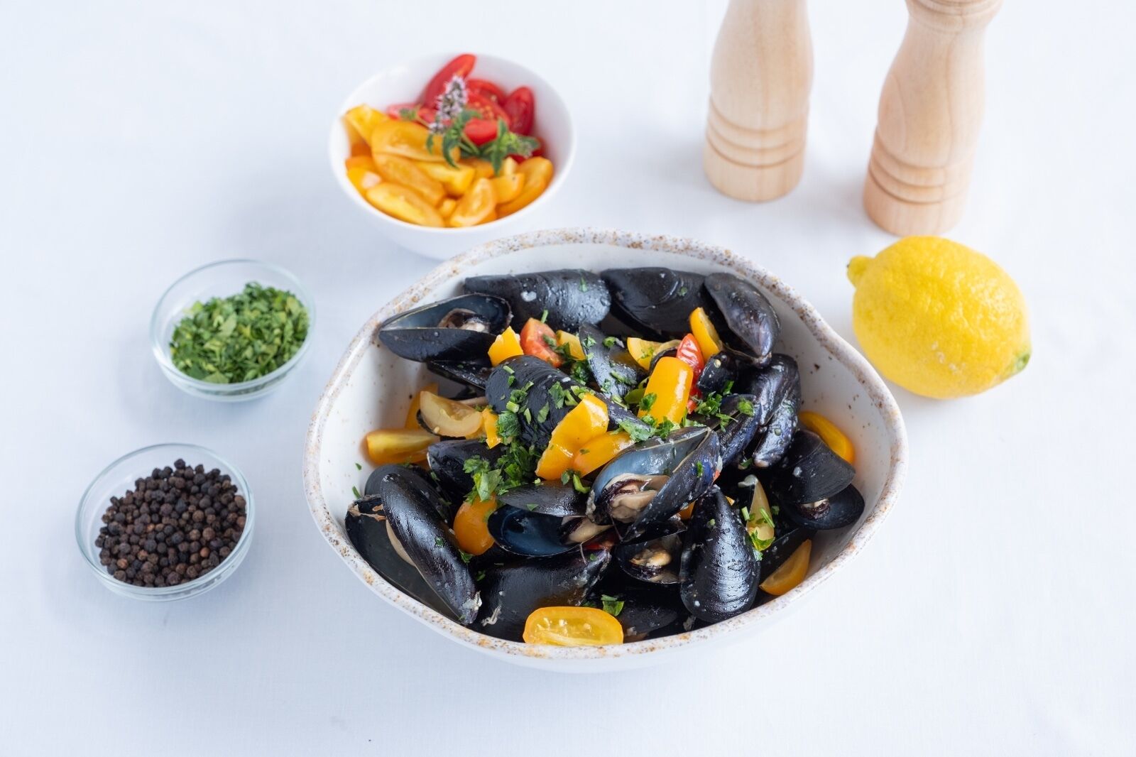 Una ciotola di cozze con verdure e erbe aromatiche, circondata da piccole ciotole di ingredienti e un limone.