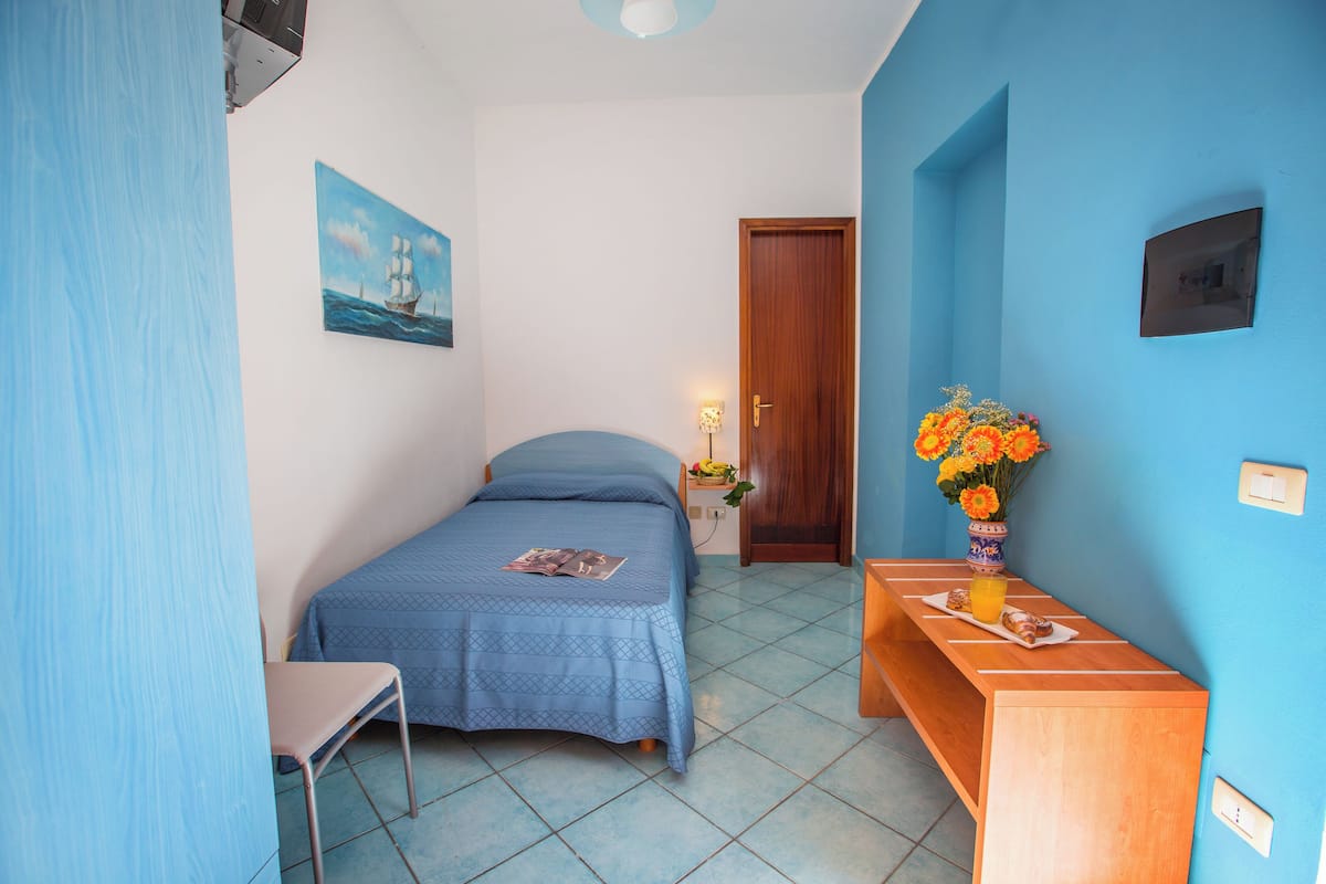 Un dormitorio con cama, escritorio, silla, un cuadro y un florero con flores.