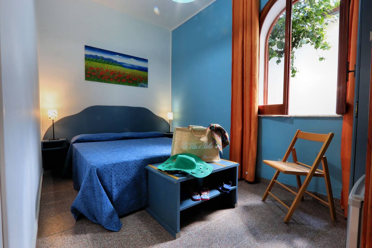 Un dormitorio con cama, mesita de noche, silla, ventana y un cuadro en la pared.