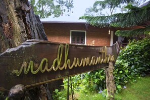 A wooden sign with the name 'Madhumathi' in cursive script, mounted on a tree trunk in a garden setting with a brick building in the background. - Madhumanthra Resort (Devikolam)