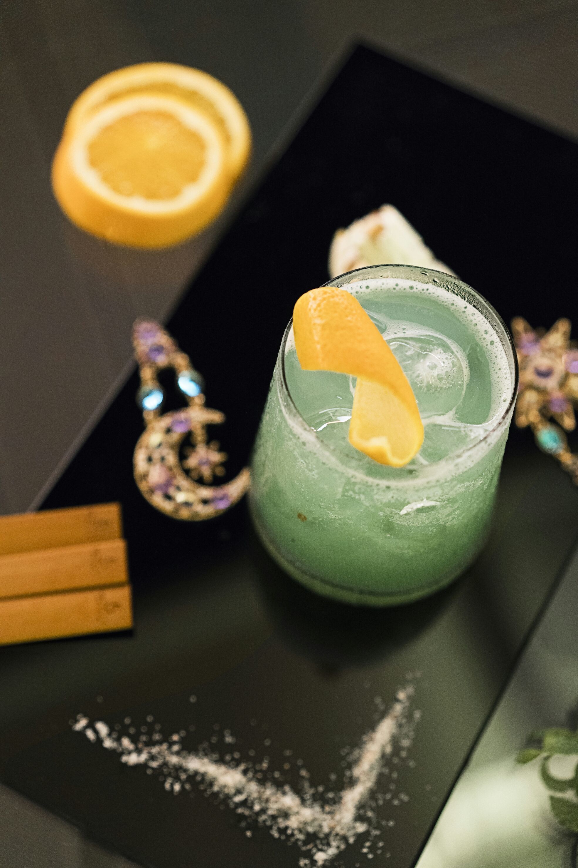 A green cocktail with an orange garnish, a crescent moon-shaped earring, and a small orange cube on a black tray.