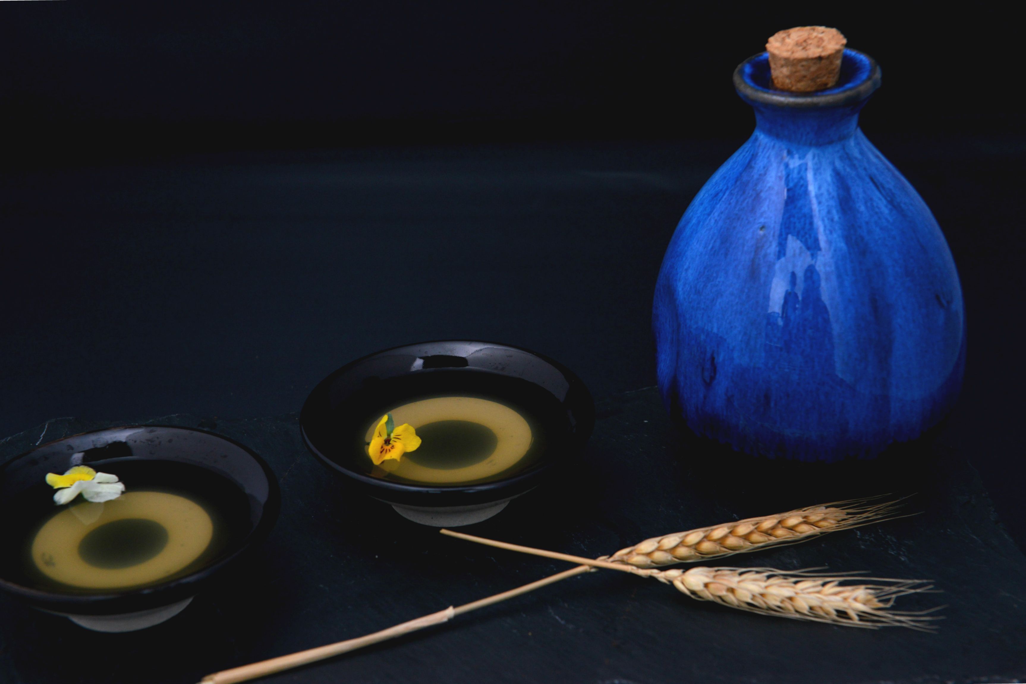 Un fond sombre sur lequel on aperçoit un vase bleu, deux bols noirs contenant chacun une fleur, une fleur jaune et deux tiges de blé.