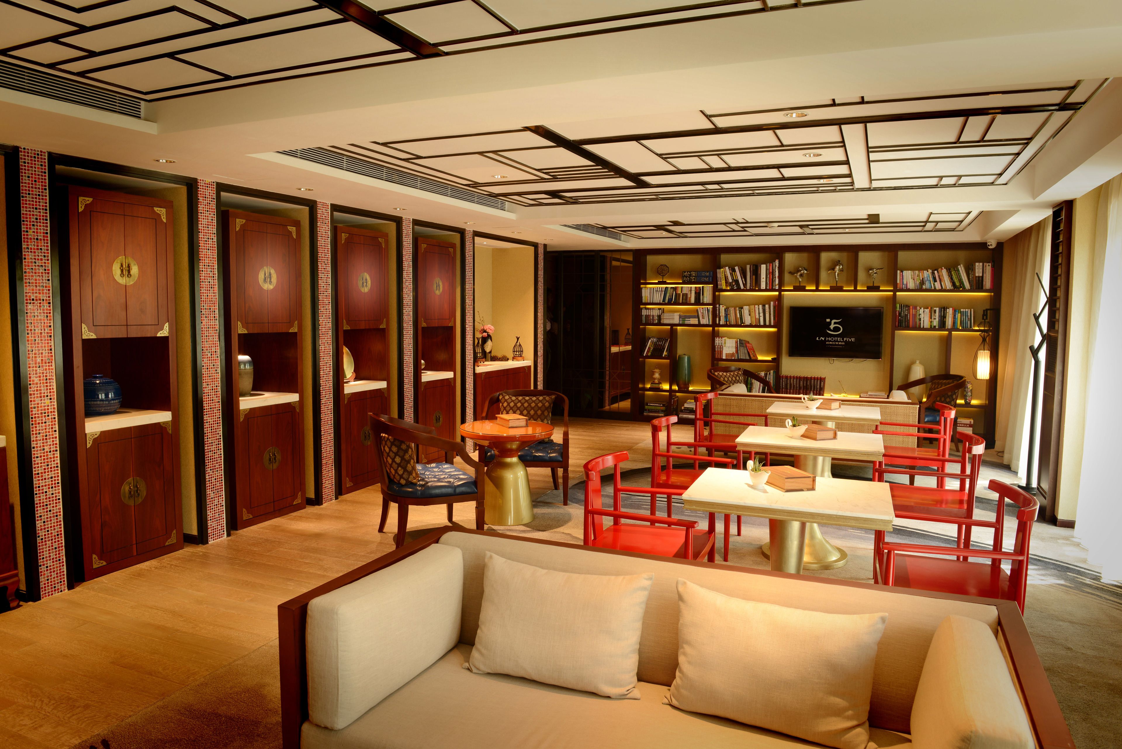 Un hall d’hôtel avec un canapé, des fauteuils rouges, une table, une bibliothèque et un écran de télévision.