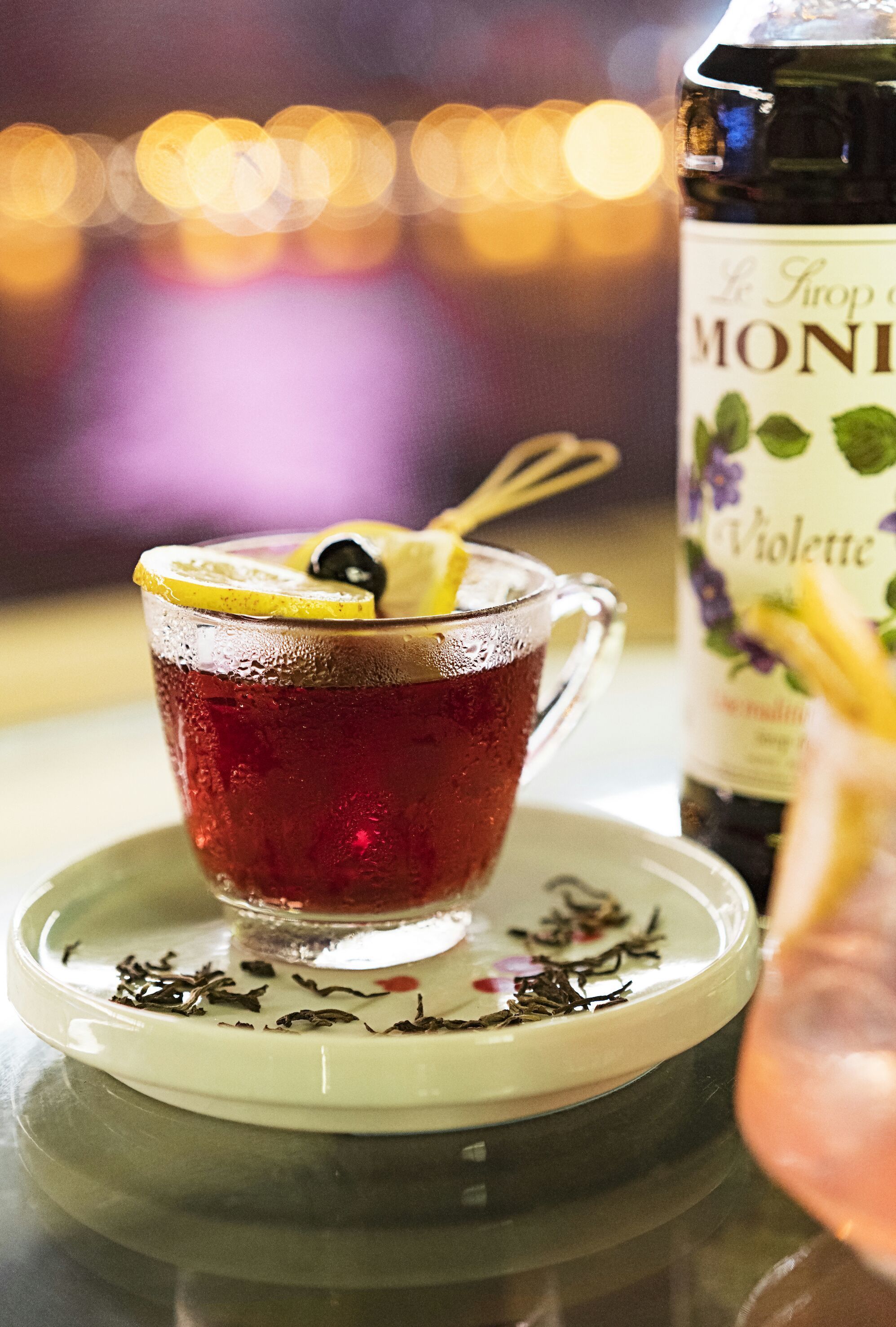 A glass of red beverage with a lemon slice and a spoon, a bottle of 'Le Sirop de Moni' on a saucer with tea leaves.