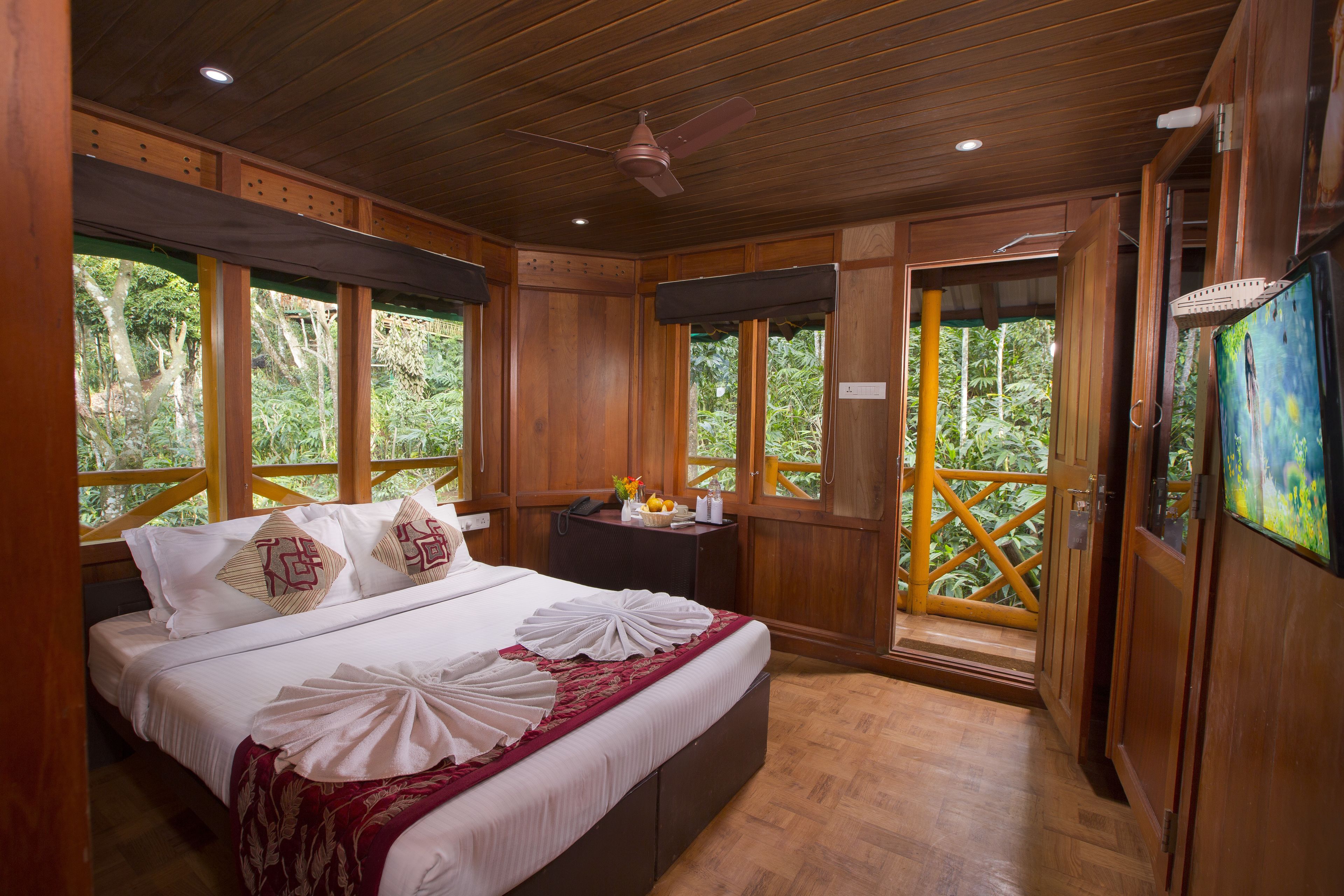 A wooden room with a large bed, wooden ceiling, and windows overlooking a lush garden.
