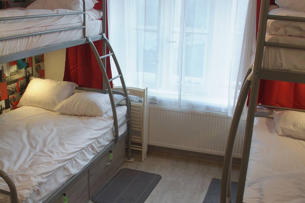 A dormitory room with bunk beds, a radiator, a window with curtains, and a wall decorated with photos.