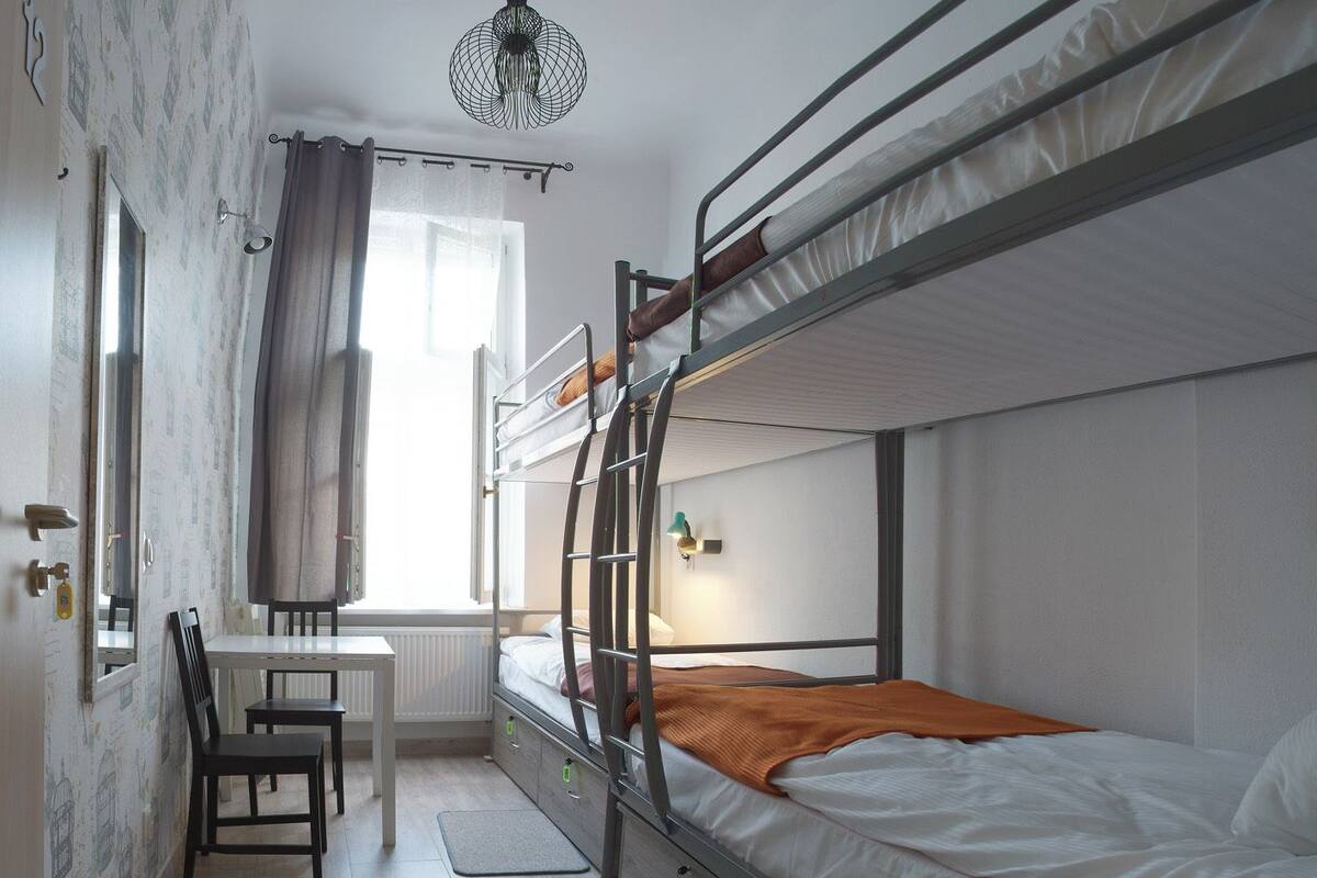 A dormitory room with bunk beds, a desk, a chair, a radiator, and a window with curtains.