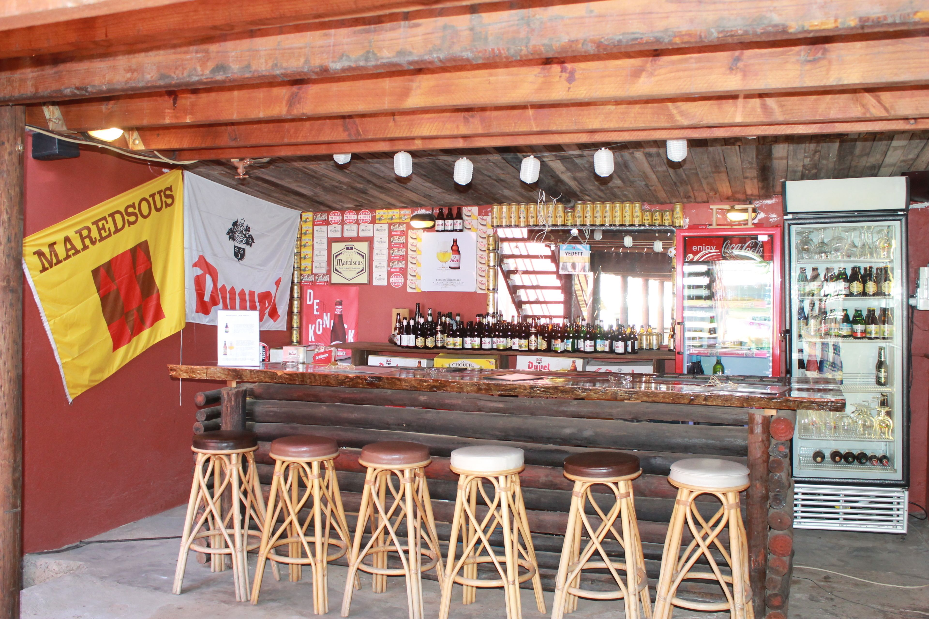 Un bar con taburetes de madera, un refrigerador lleno de bebidas y varios carteles en la pared.