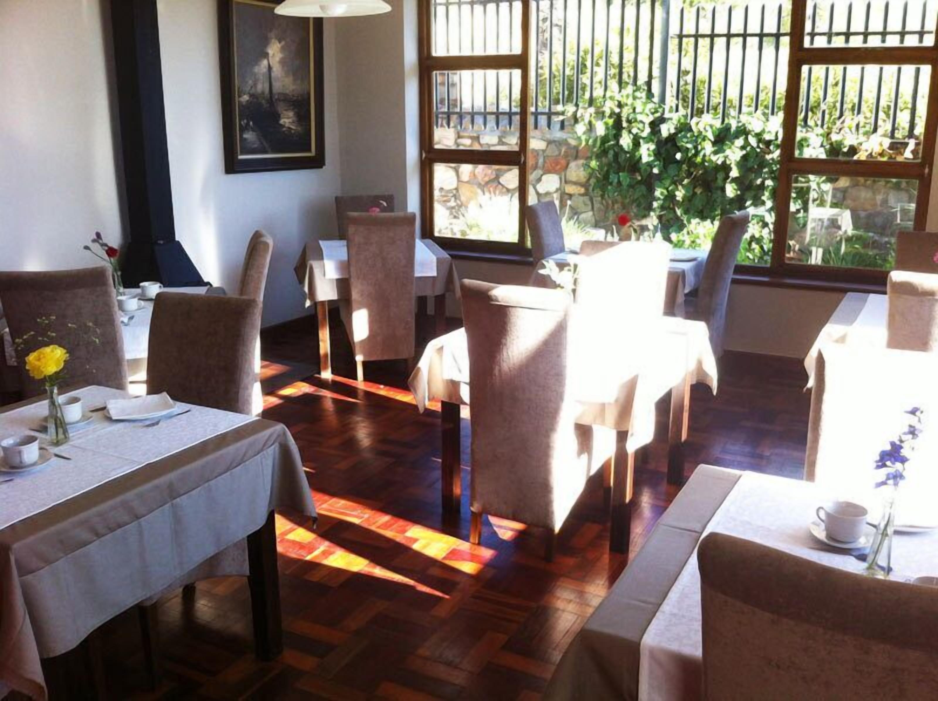 A dining area with multiple tables and chairs, large windows with a view of greenery, and a painting on the wall.
