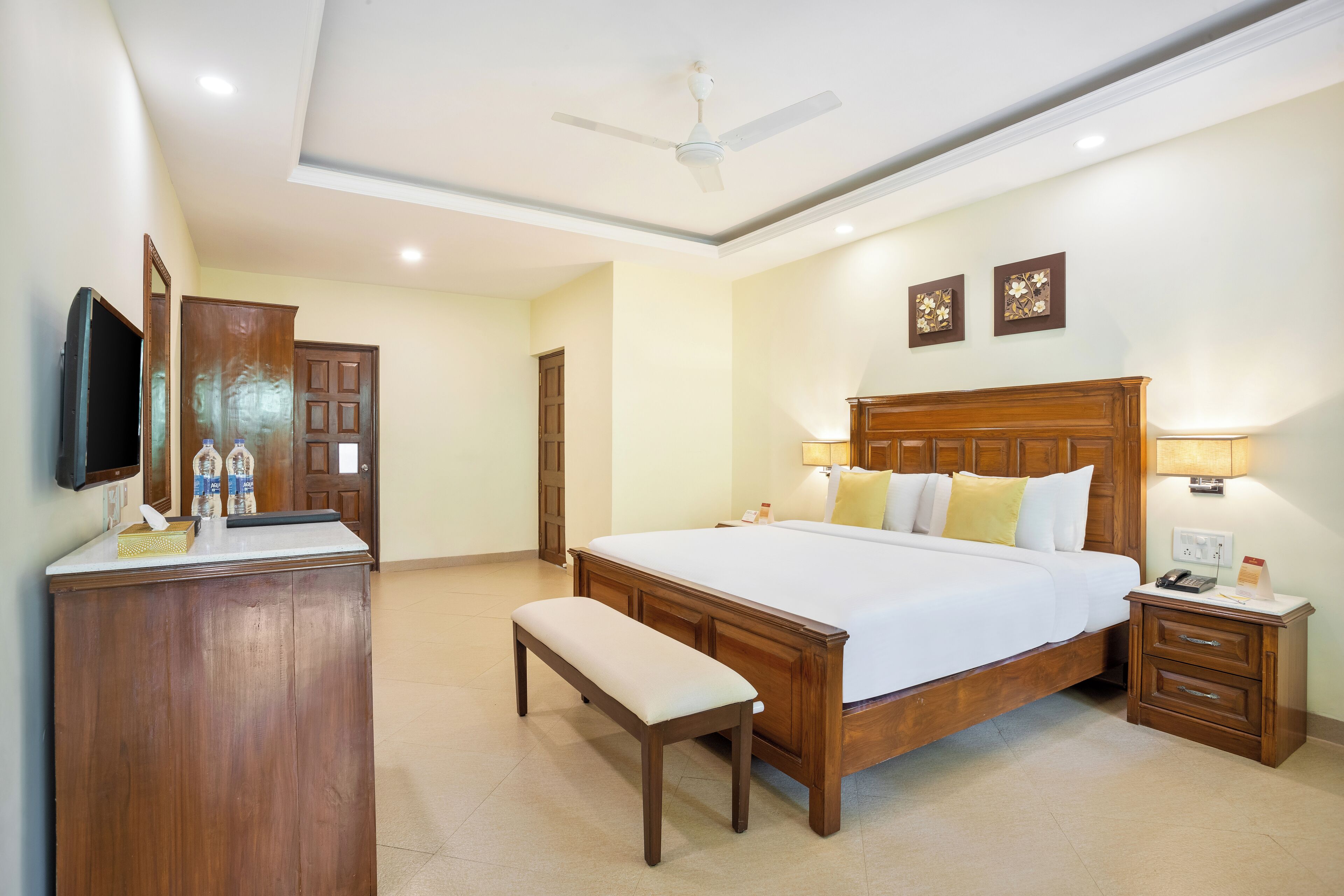 A hotel room with a large bed, bedside tables, a bench, a desk with a flat-screen TV, and a ceiling fan.