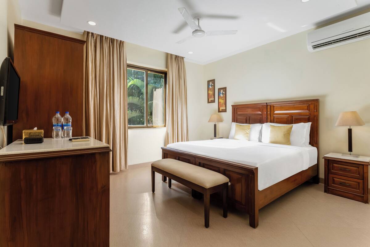 A hotel room with a large bed, a wooden desk, a bench, two bedside lamps, and an air conditioning unit.
