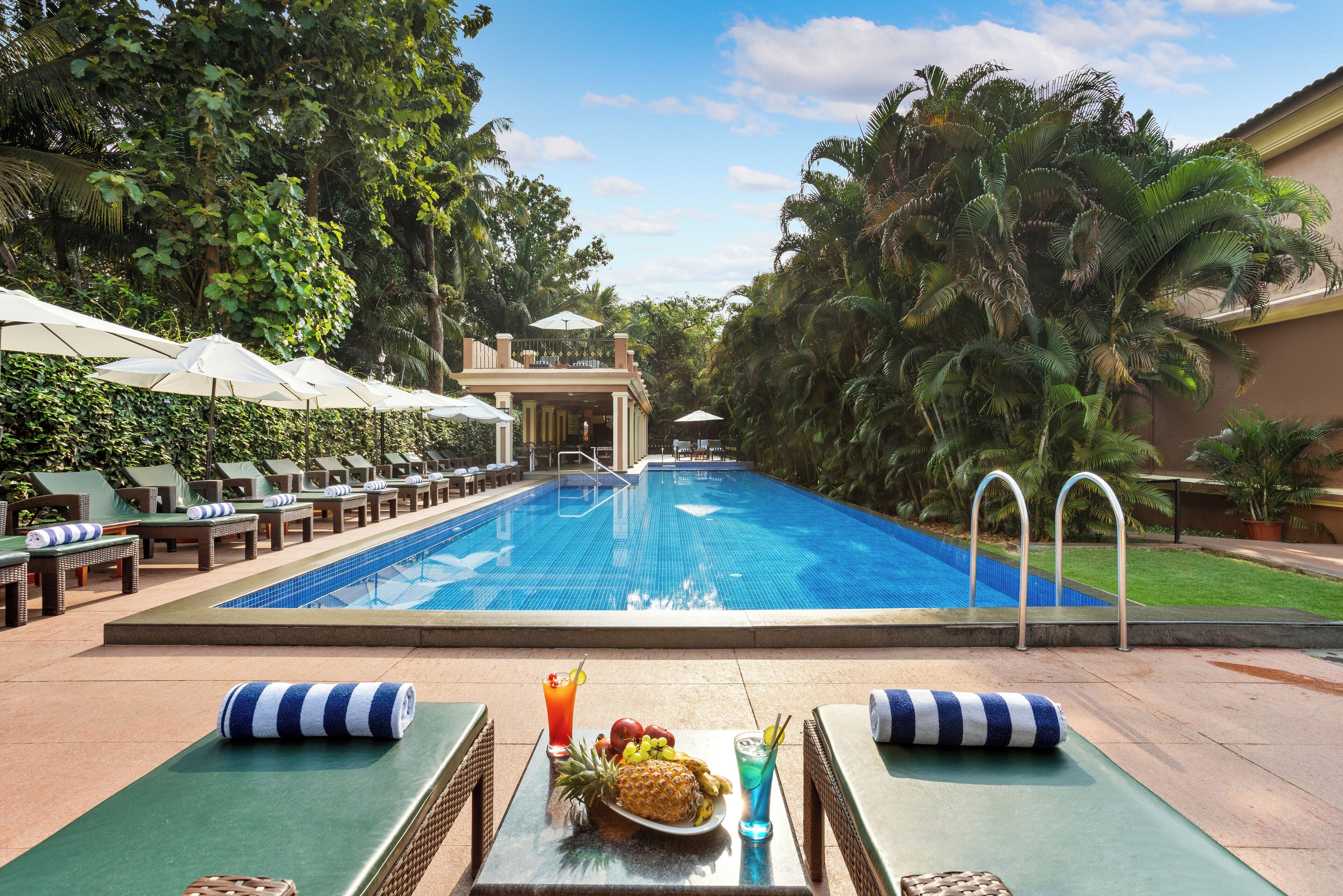 Zona de piscina con sillones de descanso, sombrillas y una mesa con una bandeja de frutas y bebidas.