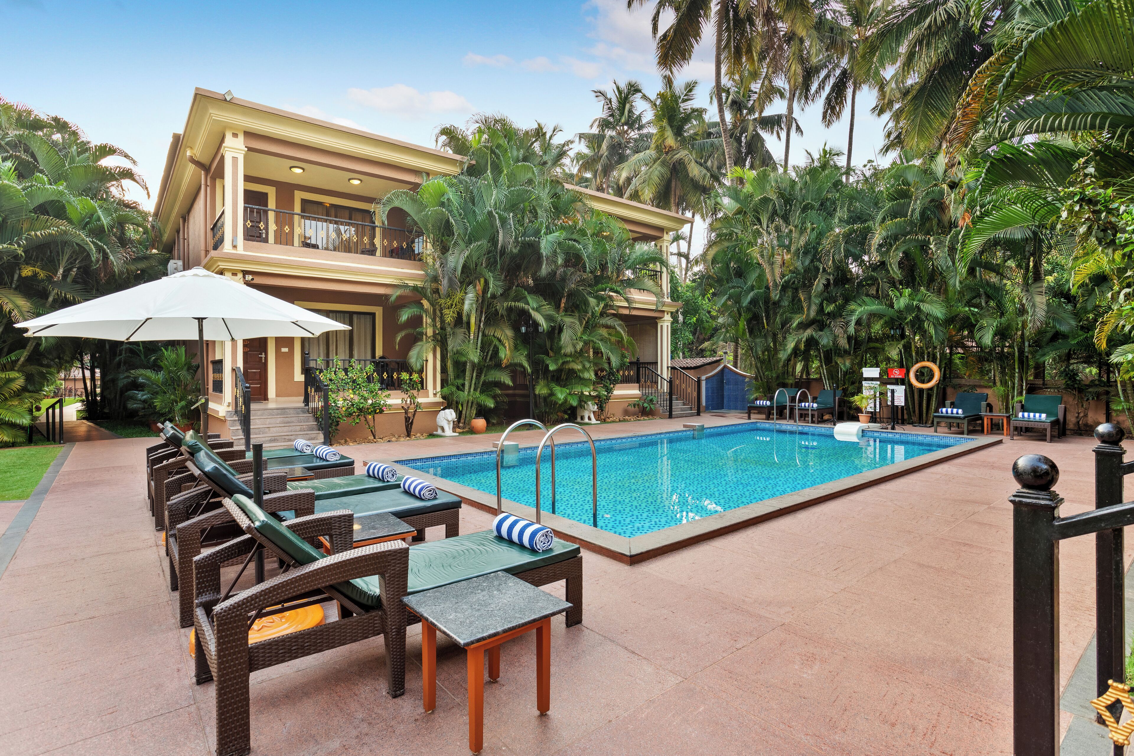 Zona junto a la piscina con sillones de descanso y una piscina, rodeada de palmeras y un edificio de dos pisos.