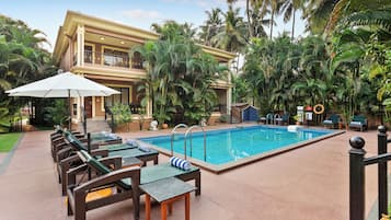 Zona junto a la piscina con sillones de descanso y una piscina, rodeada de palmeras y un edificio de dos pisos.