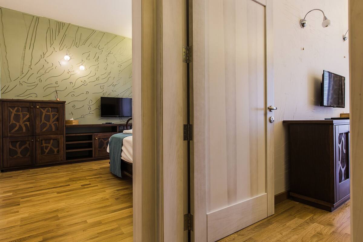 Quarto de hotel com piso de madeira, uma cama, televisão, guarda-roupa e parede com um mural de árvore.
