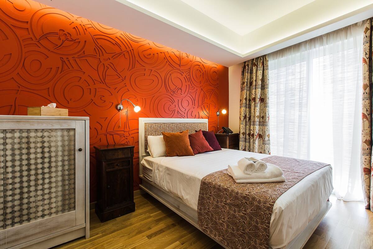 Quarto de hotel moderno com uma cama grande, uma mesa de cabeceira, uma luminária e uma janela com cortinas.