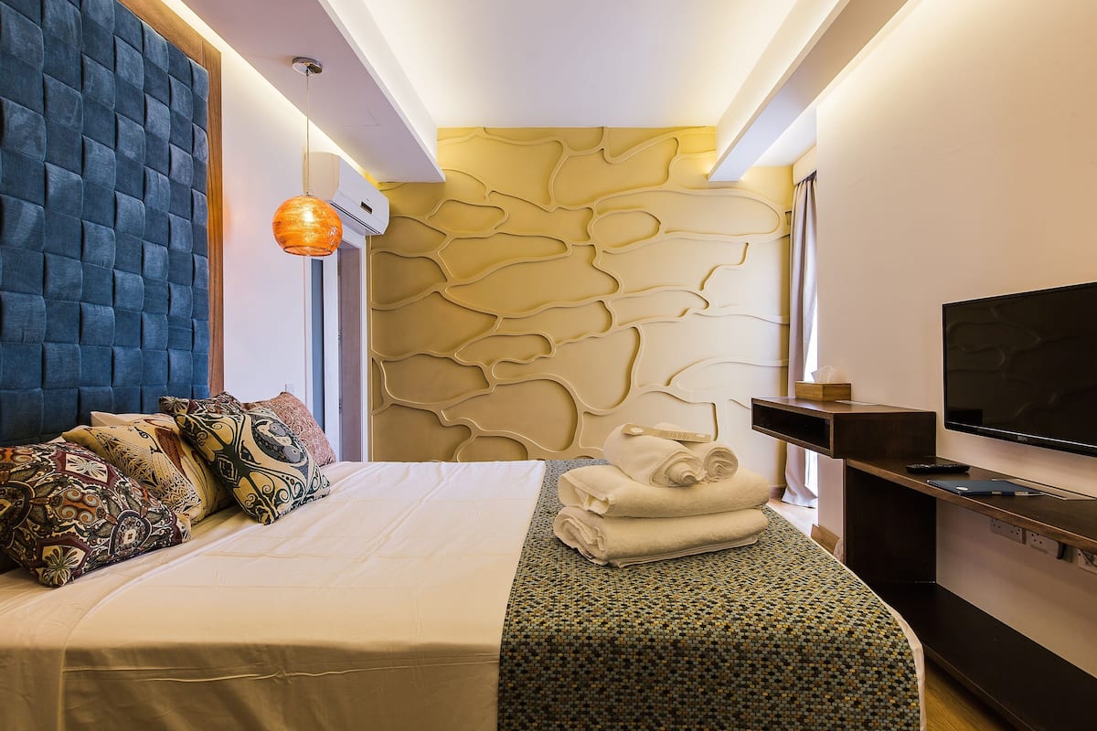 Quarto de hotel com uma cama, televisão, parede com textura única e luminária no teto.