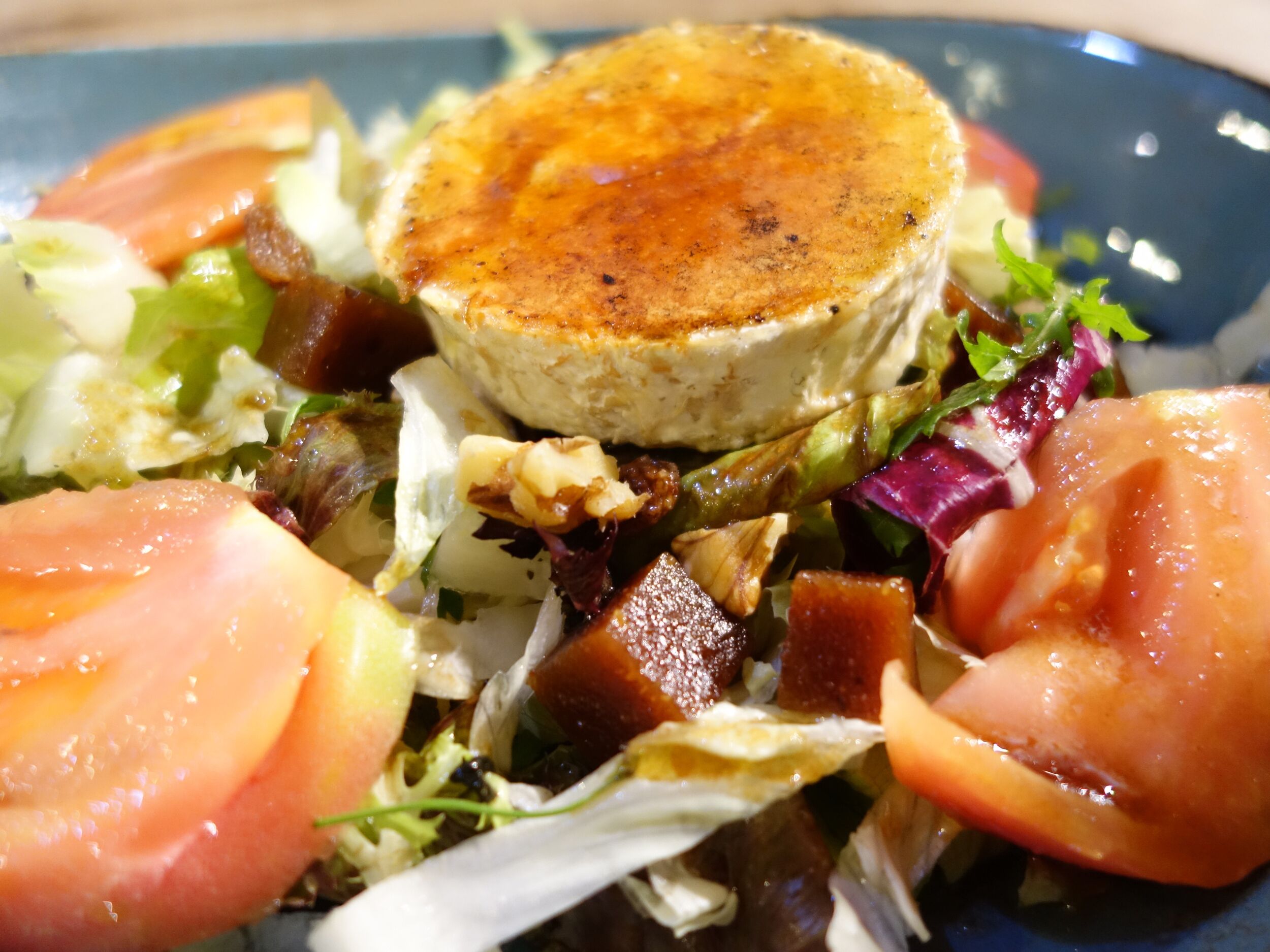 Un plato con un elemento redondo y tostado, rodeado de hojas verdes variadas y rodajas de tomate.