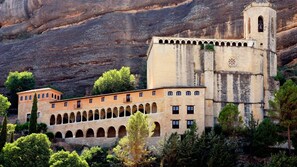 Um edifício histórico com uma torre de sino e várias arcadas, rodeado por árvores e falésias rochosas.