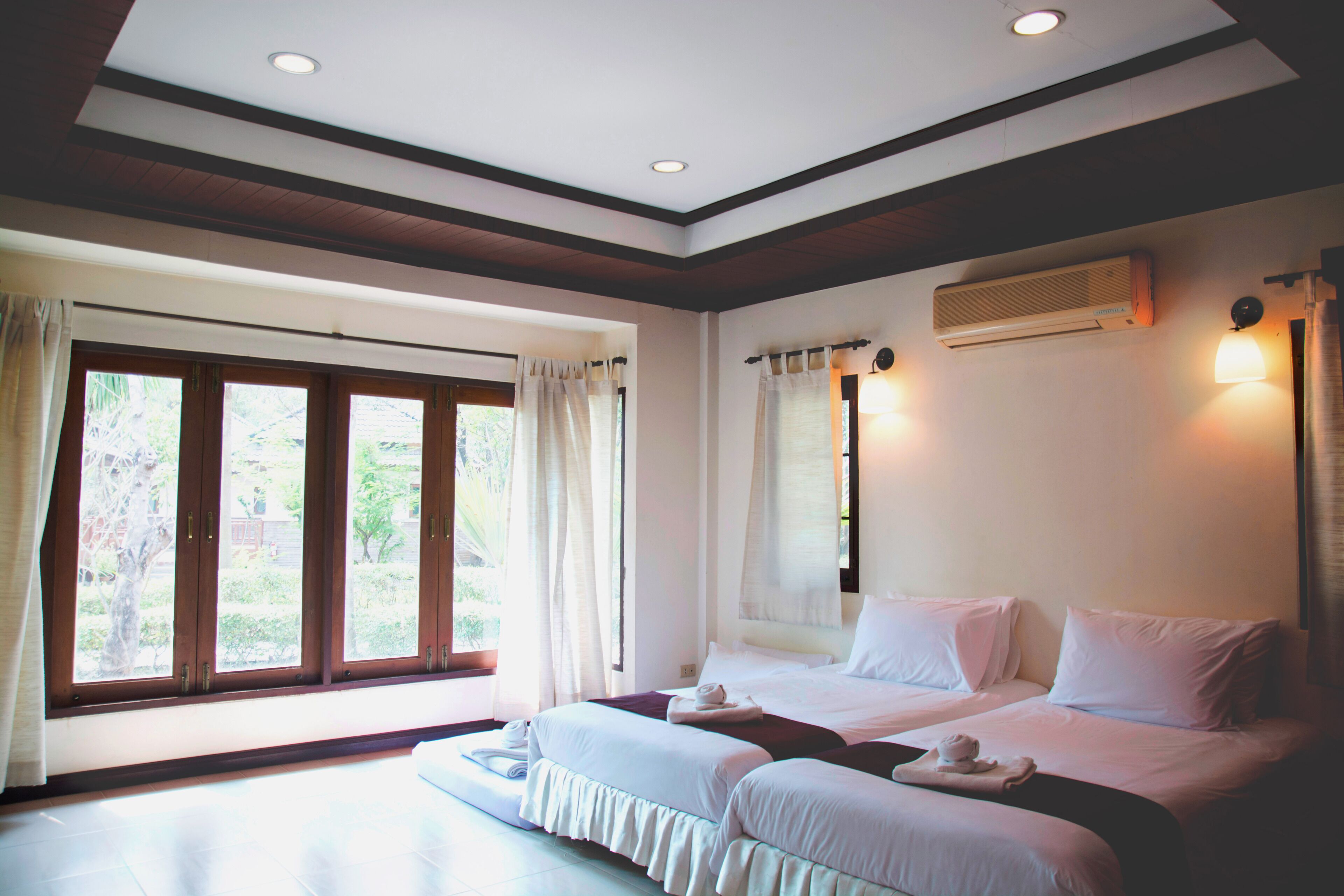 A hotel room with two beds, a large window with curtains, an air conditioning unit, and a ceiling light.
