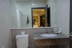 Une salle de bain avec des toilettes blanches, un lavabo doté d’un plan de travail en granit et un miroir qui reflète une pièce comprenant un lit et une porte.