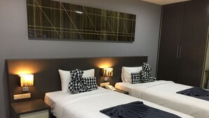 Habitación de hotel con dos camas, cada una con una almohada y una lámpara de noche. Se observa un armario y un aplique en la pared sobre las camas.