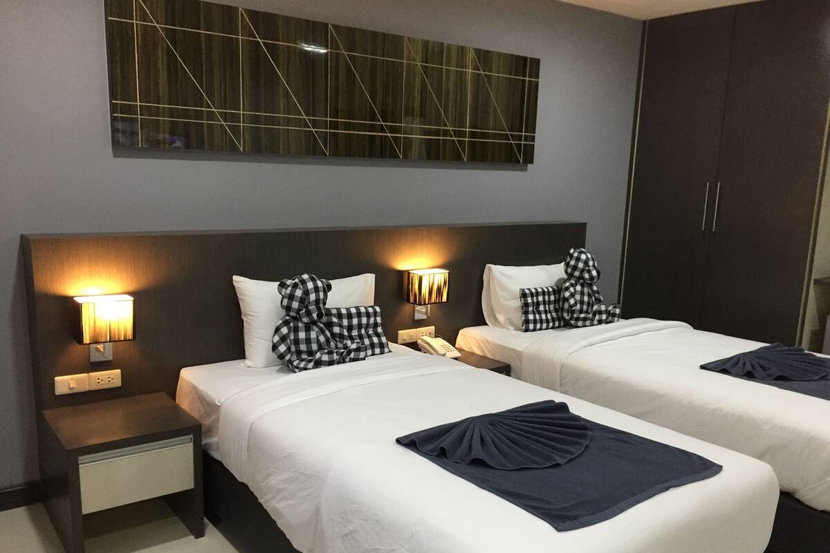 A hotel room with two beds, each with a pillow and a bedside lamp. There is a wardrobe and a wall-mounted light fixture above the beds.