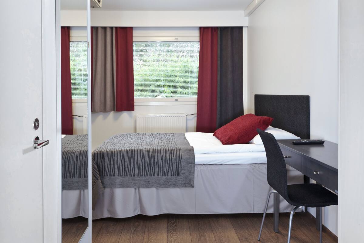 A bedroom with a bed, a desk, a chair, and a window with curtains.