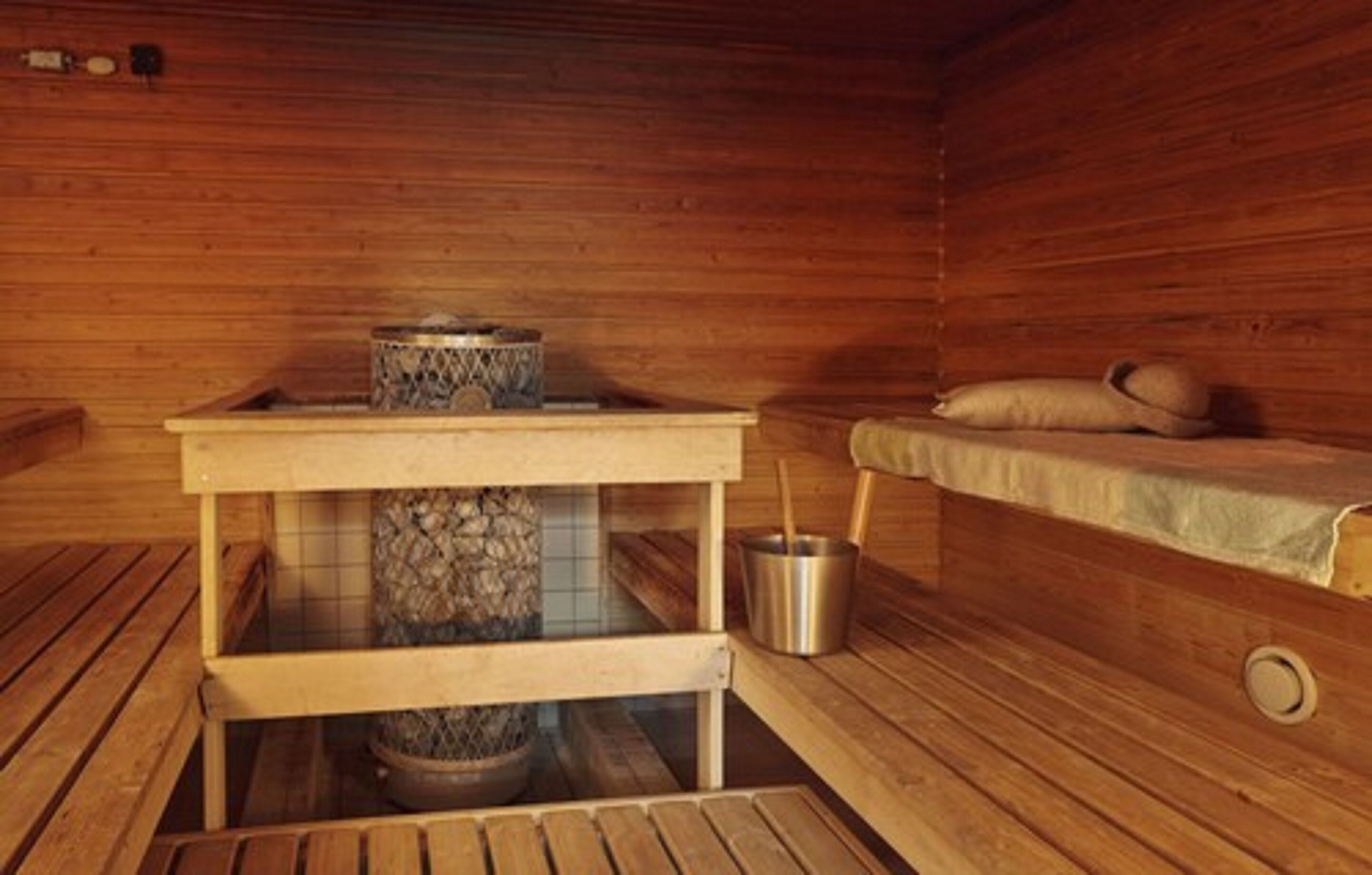 A wooden sauna room with a stone heater, a metal bucket, and a bench with a cushion.