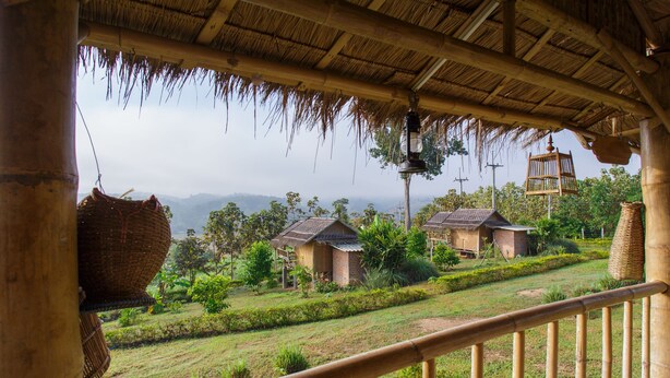 Estructura con techo de paja y baranda de bambú, con vista a un paisaje rural con casas pequeñas y vegetación.