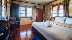 Habitación de hotel con una cama, un escritorio y una silla. Hay dos ventanas con cortinas, una puerta y un ventilador de pared.