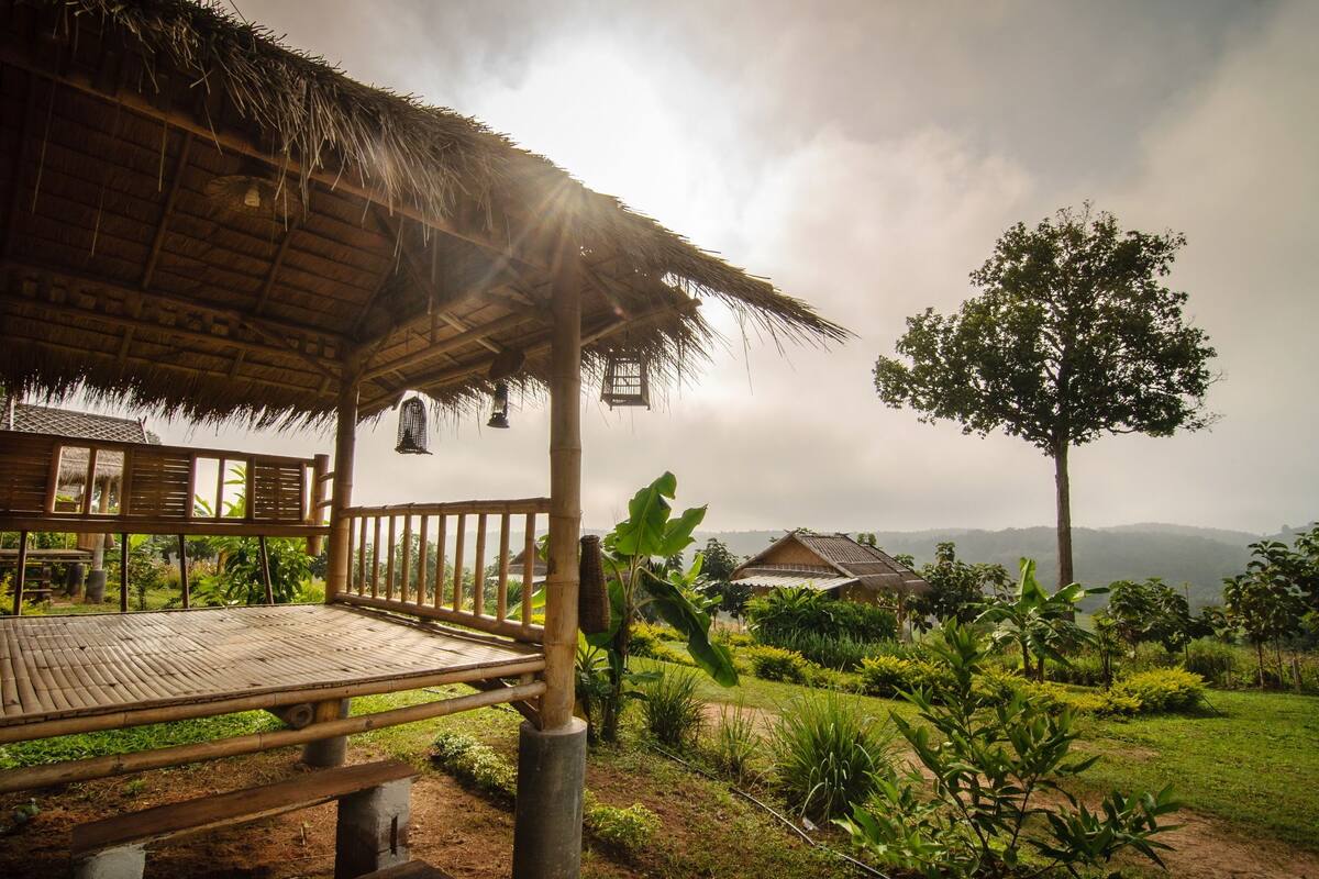 A bamboo structure with a thatched roof, a wooden bench, and a view of a garden with various plants and a distant mountain.