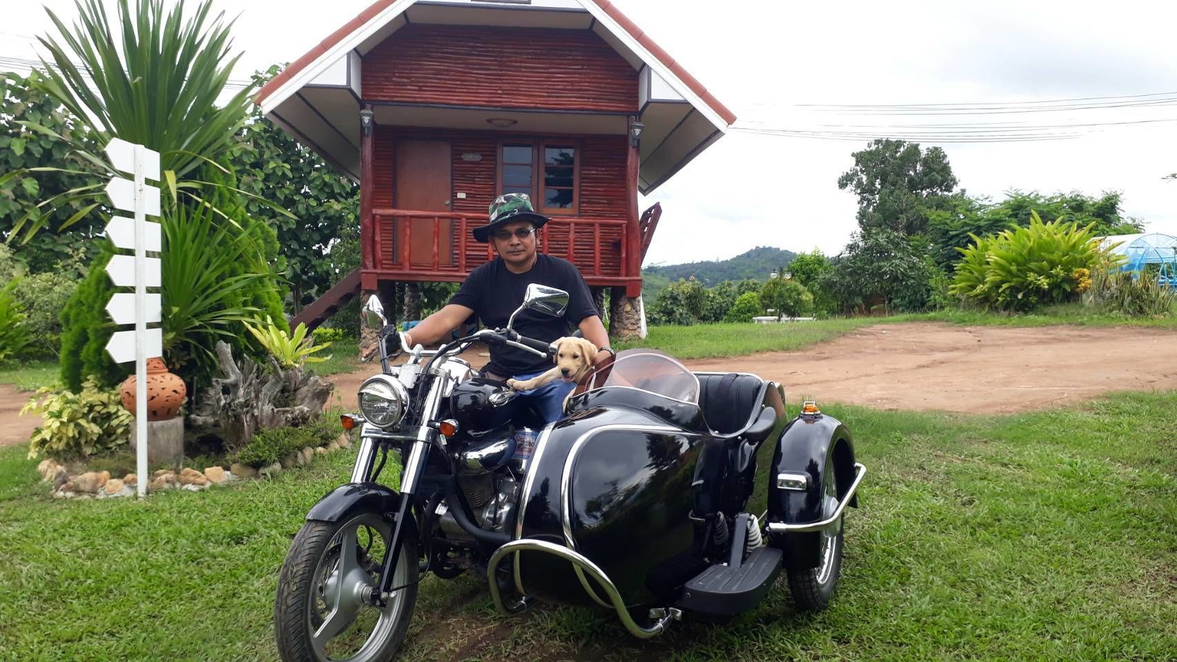 A person sitting on a motorcycle with a sidecar, parked in front of a house with a red roof and a signpost with multiple arrows.