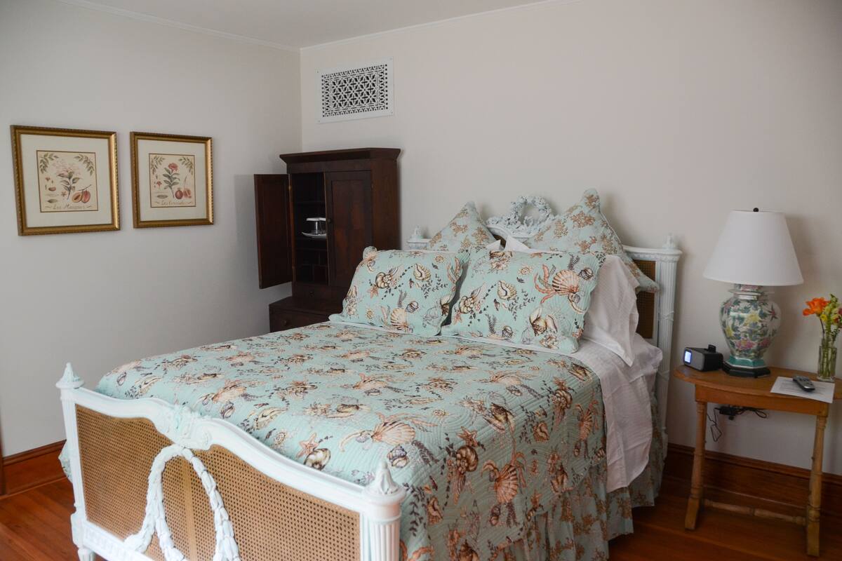 A bedroom with a bed, a nightstand, a lamp, and two framed botanical prints on the wall.