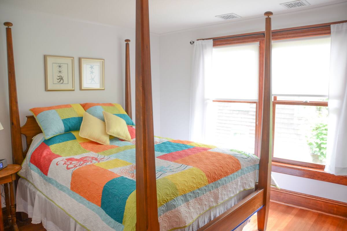 A four-poster bed with a colorful quilt, wooden headboard, and a nightstand with a lamp.