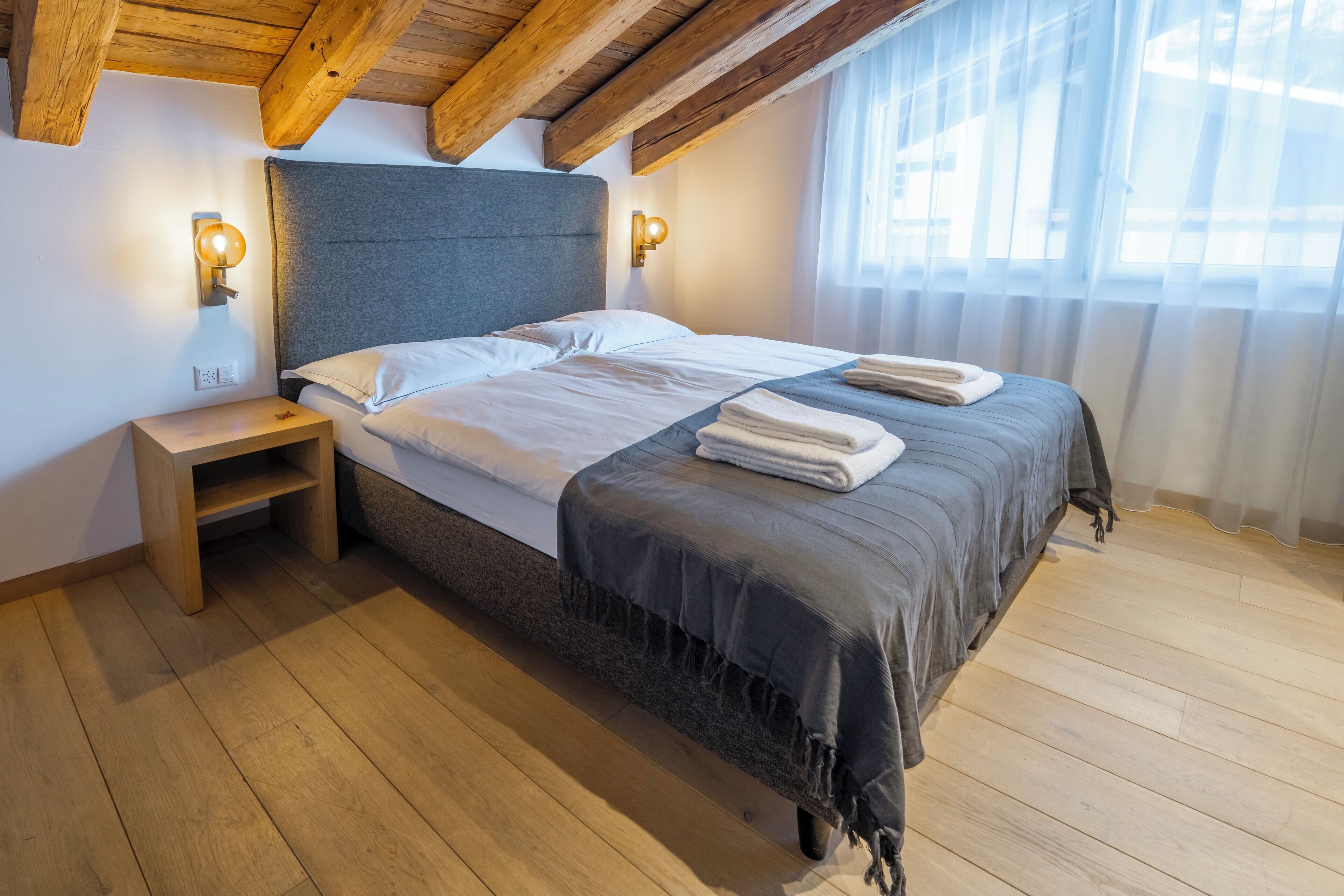 A bedroom with a wooden ceiling, a bed with a grey headboard, two bedside lamps, and a window with sheer curtains.